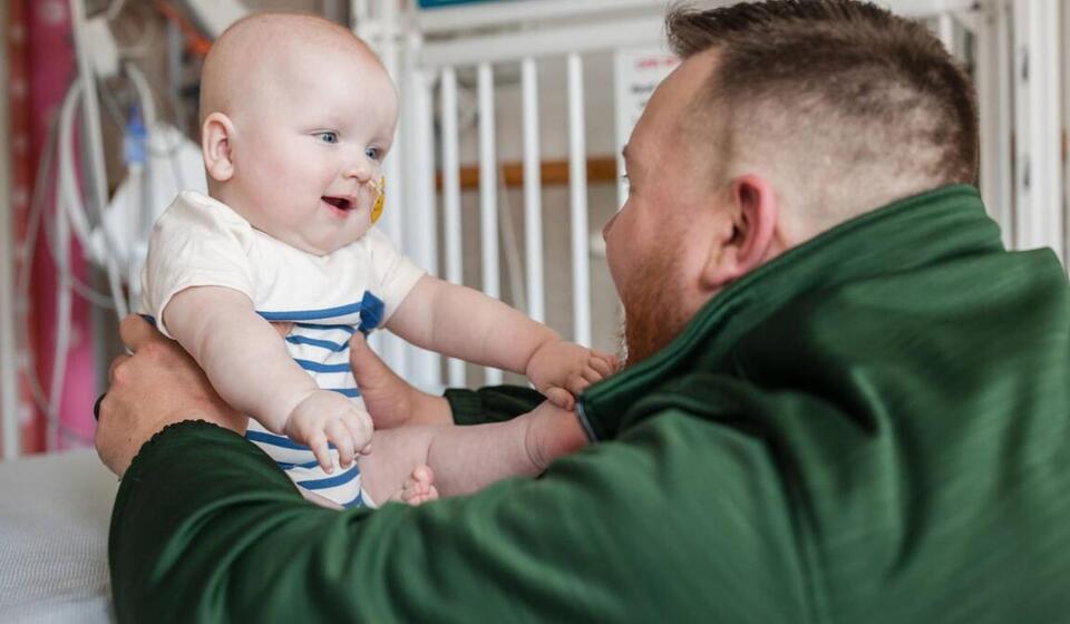 A man in a green jacket looks at a baby in front of him, both looking excitedly at  each other